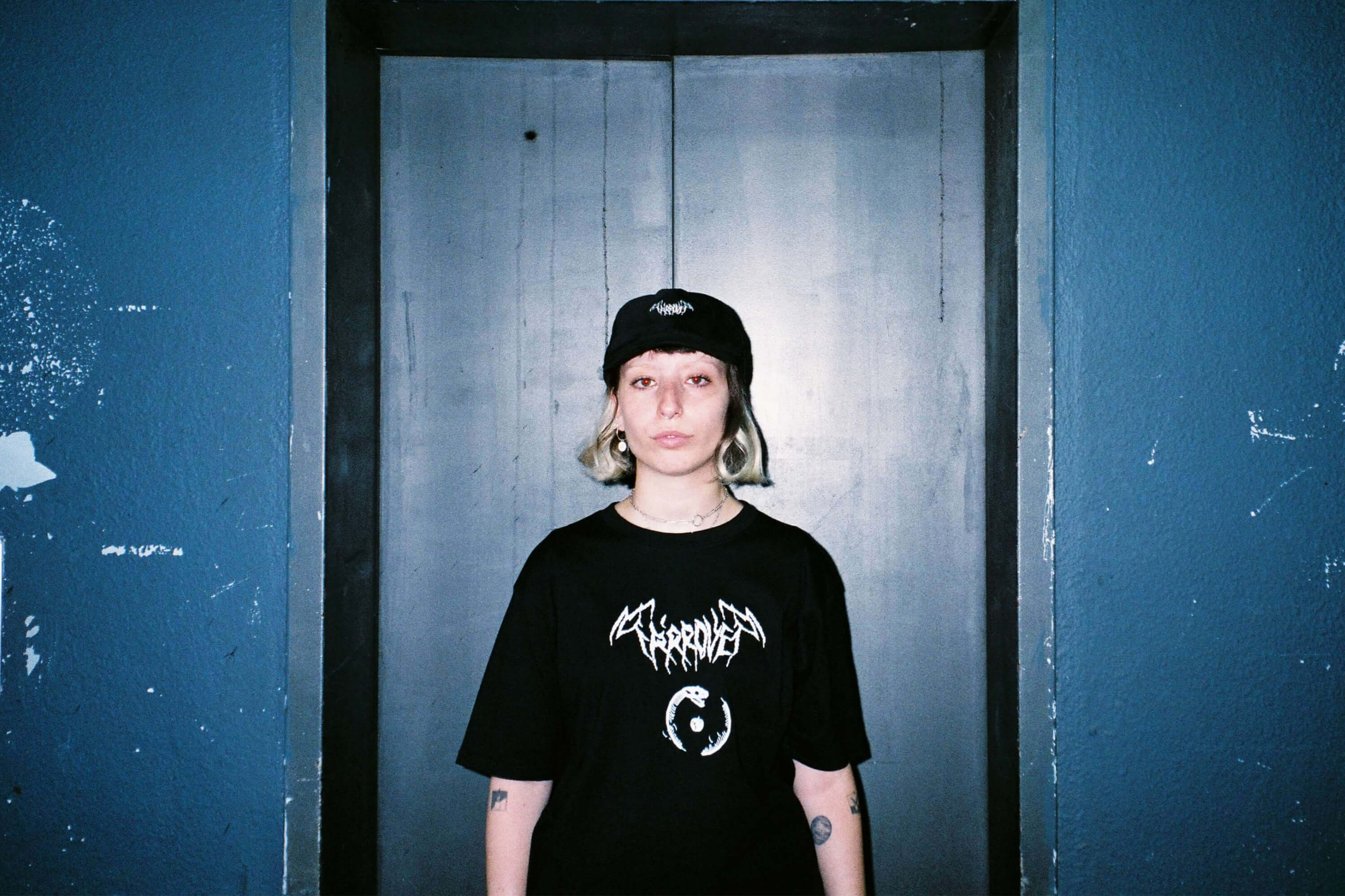 A person with short, light blonde hair stands facing the camera in front of a closed metal elevator door. They wear a black cap and oversized black T-shirt, both displaying the “Ta’arovet” logo in sharp, gothic-style white lettering. The wall behind them is dark teal with visible scuffs and peeling paint, reinforcing a gritty, urban streetwear aesthetic. The person maintains a neutral expression, contributing to the image’s somber and defiant tone.
